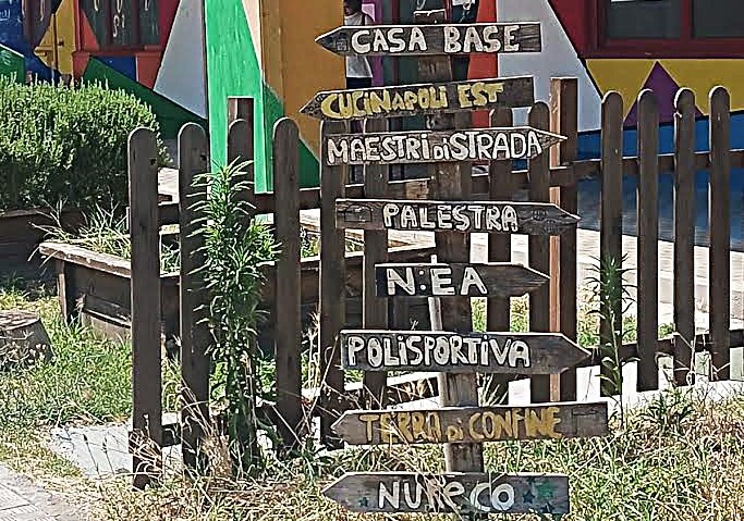 Cartelli in legno indicano le attività del Centro Polifunzionale Ciro Colonna a Ponticelli: Casa Base, Cucina Napoli Est, Maestri di Strada, palestra, polisportiva, Terra di Confine e Nureco.