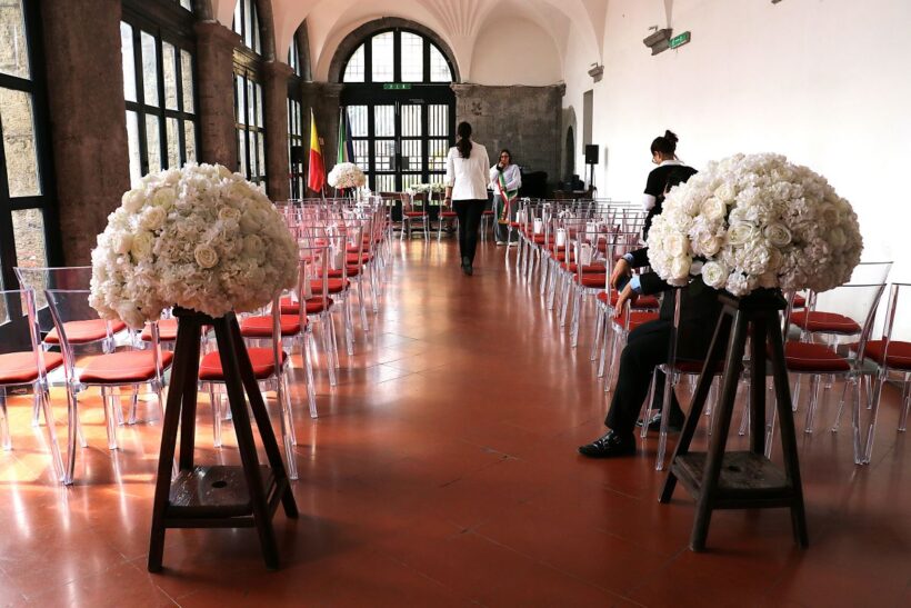 Interno della sala per le cerimonie civili a Castel Nuovo di Napoli, con file di sedie trasparenti e grandi composizioni floreali bianche disposte lungo il corridoio centrale.